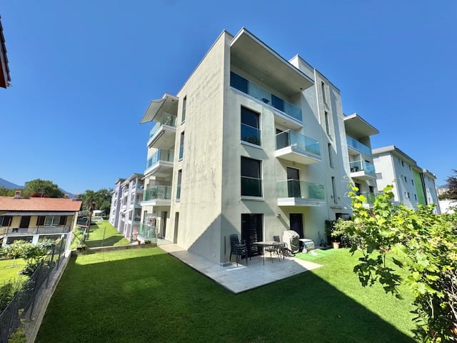 A three-story apartment building with modern design, balconies, and large windows, surrounded by a well-maintained lawn and lush greenery, creating a peaceful and picturesque environment.
