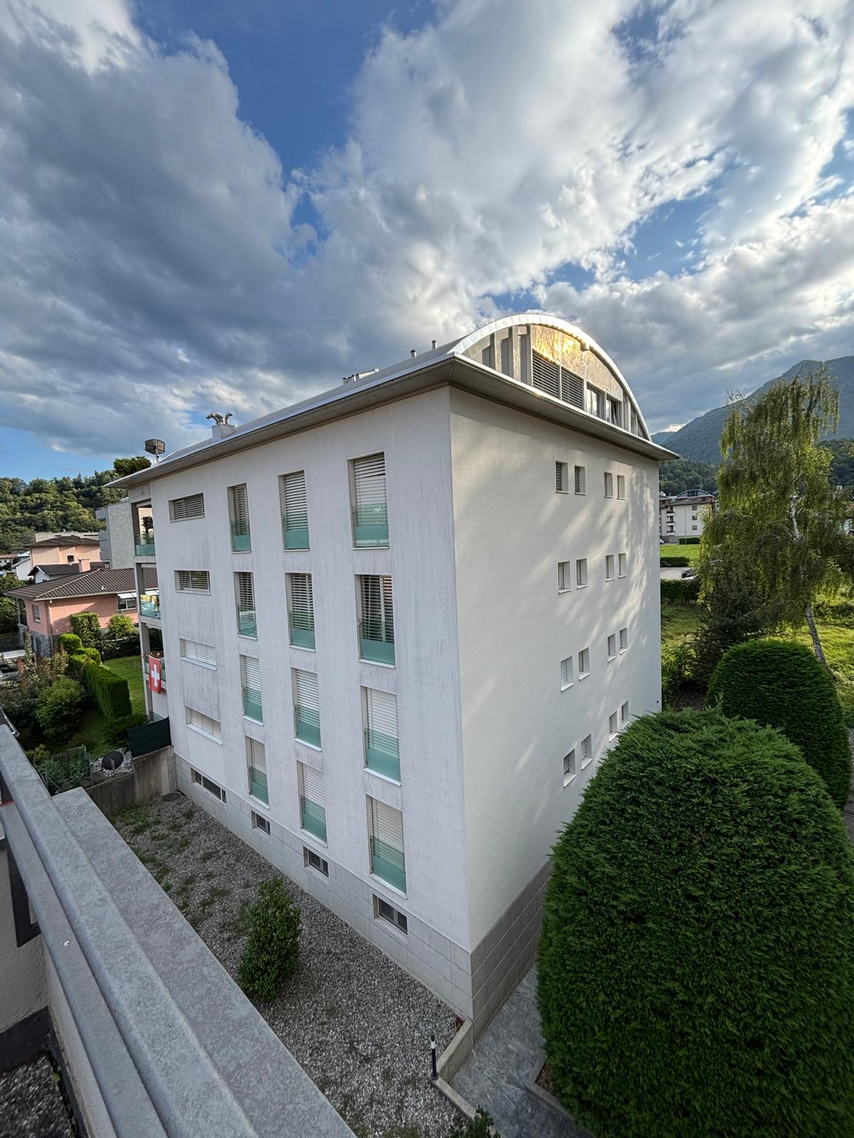 Multistory white building with multiple windows and balconies, flat roof, surrounded by greenery and mountains in the background