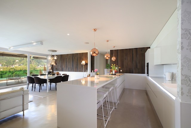 modern interior, white countertops, open-plan, large windows, large dining table, 2 stools, 2 couches, wooden wall, chandeliers