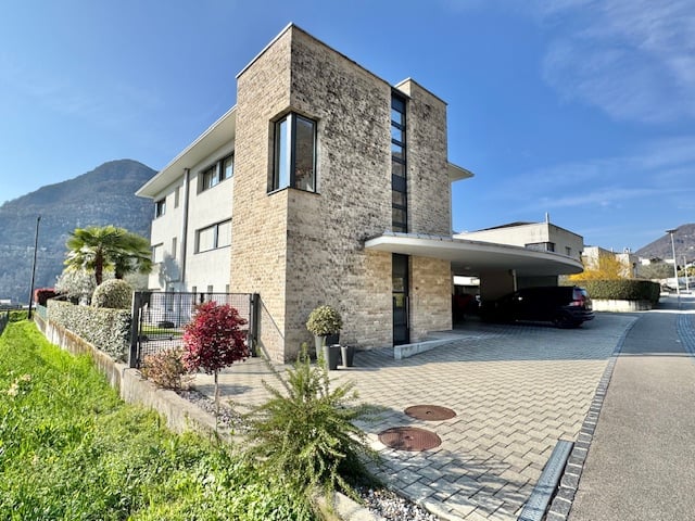 Modern two-story house with brick facade, large windows, car parked in garage, landscaped garden with plants and hedges, driveway with brick paving