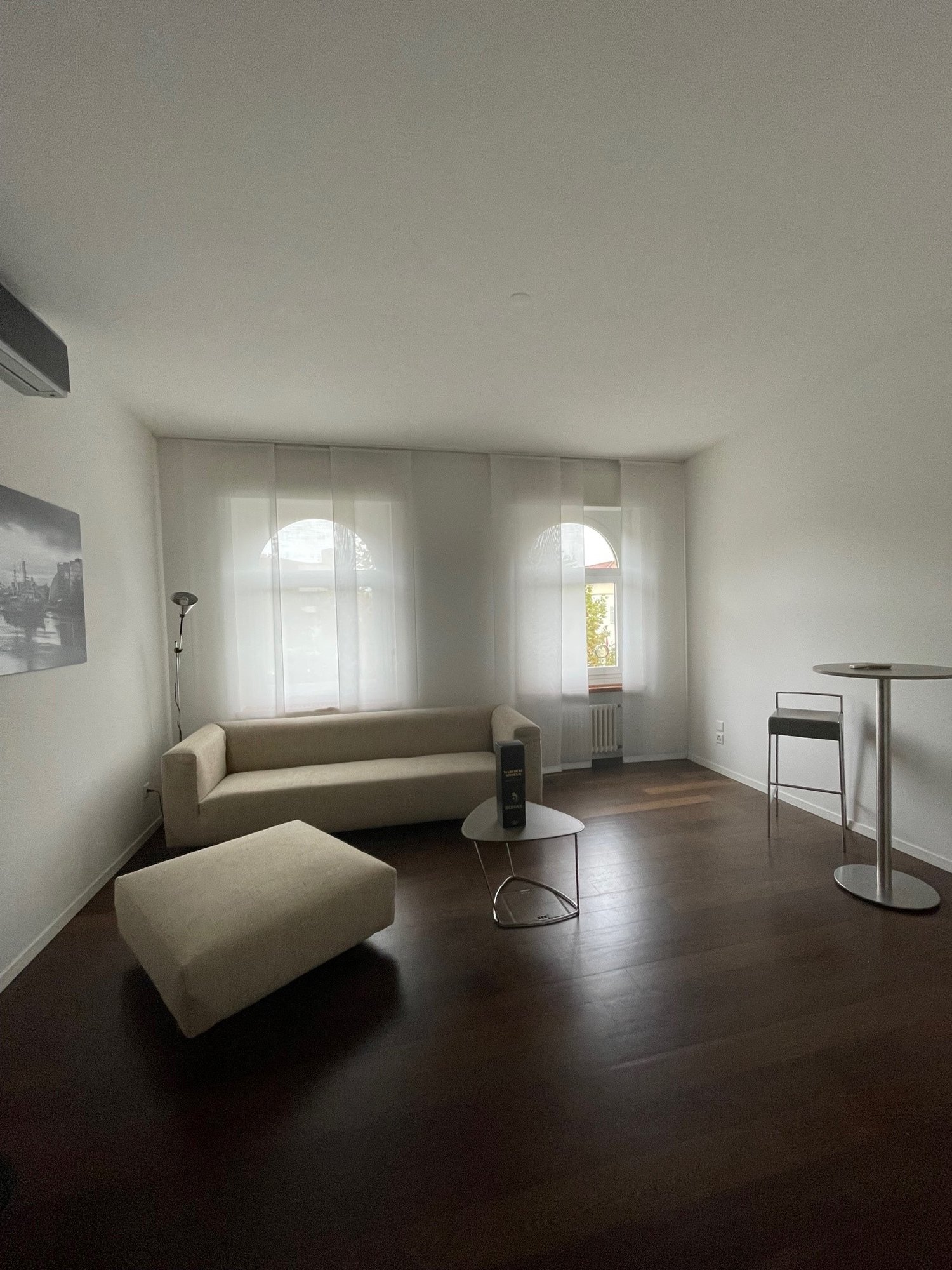 large sofa, foot stool, round table, floor lamp, two large arched windows with curtains, picture frame, hardwood floor, ceiling lamp, white walls