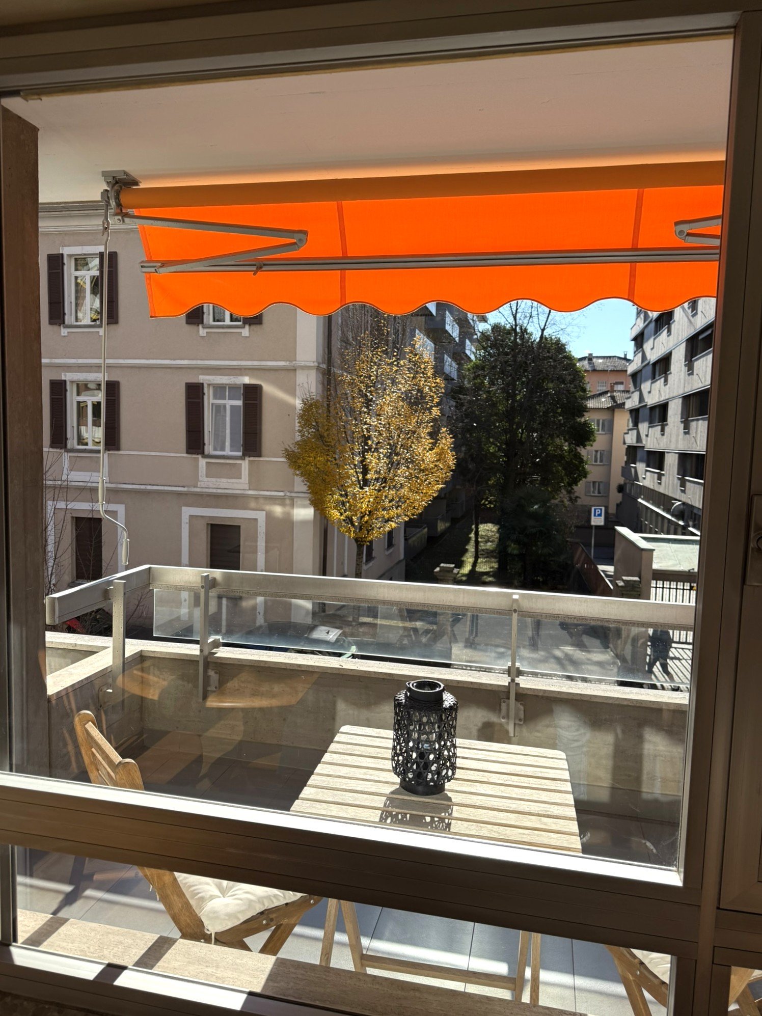 Window view of a balcony with a wooden table and chairs, metal railings, and a retractable orange awning. The view includes a tree and a street with a parked car.