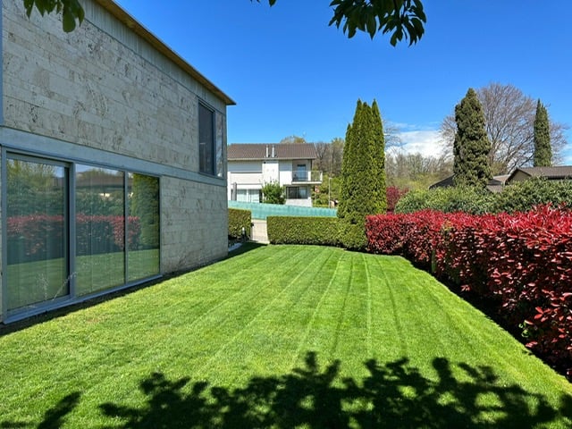 2-story modern house with large glass windows, surrounded by a well-manicured lawn, lush greenery, and trees. The property also has a parking area visible.