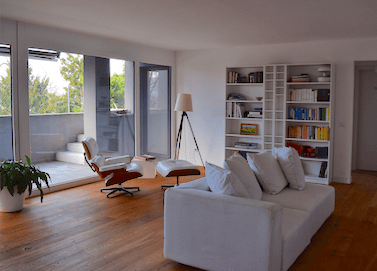 Living room, white walls, hardwood floor, glass door to terrace, white couch, armchair, books on shelf, lamp, plants