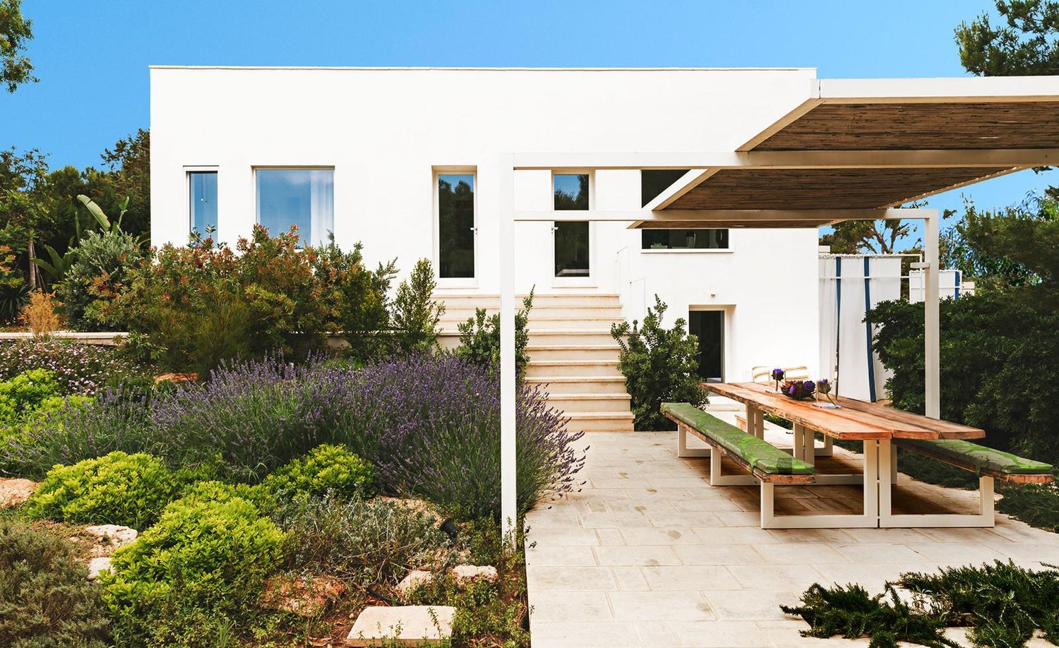 Modern white house, large windows, wooden pergola, outdoor dining area, garden with various plants