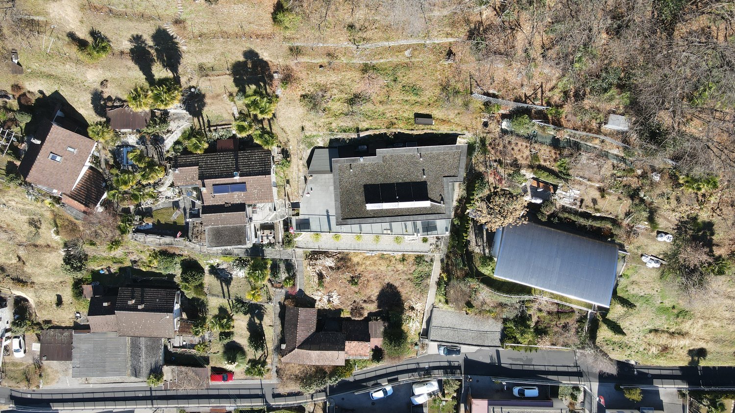 An aerial view of several houses on sloping land with grass, bushes, trees, and parked cars.