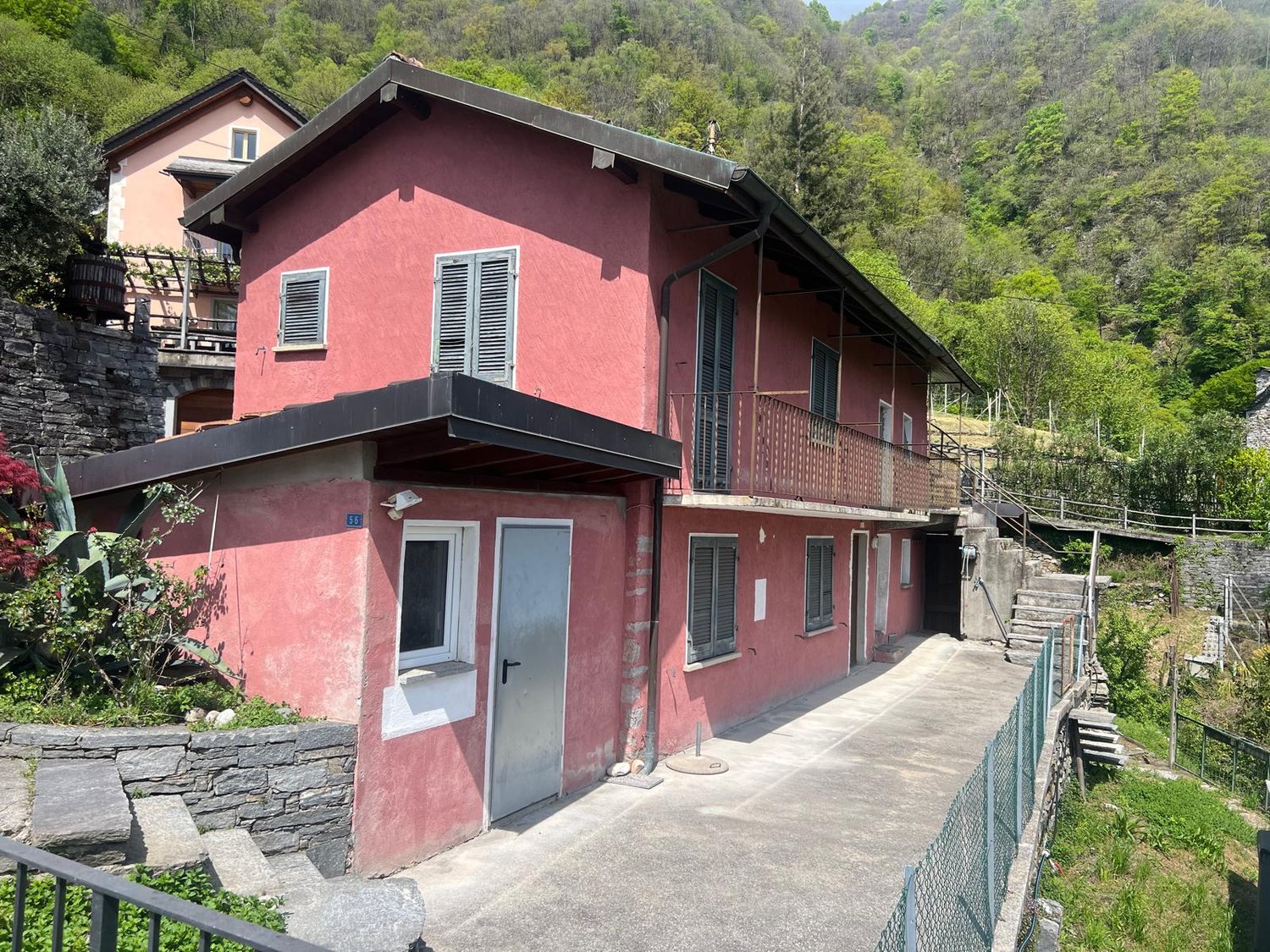 2-story pink house with a balcony, surrounded by lush greenery and a stone wall