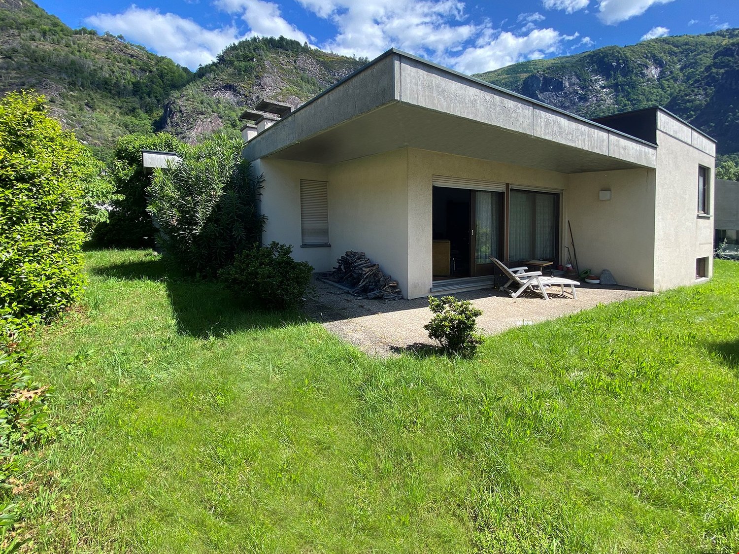 Single-story modern house with a flat roof, surrounded by lush greenery and mountains in the background. The house has large windows and a balcony or terrace area.