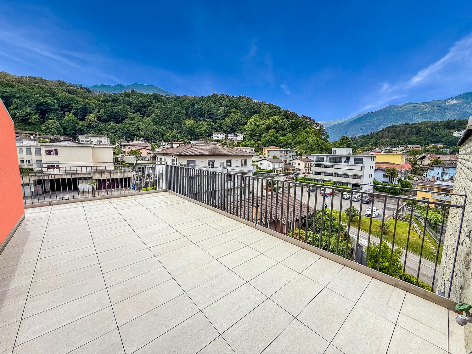 Spacious balcony with tiled flooring, metal railing, overlooking a residential area with buildings, trees, and mountains in the background.