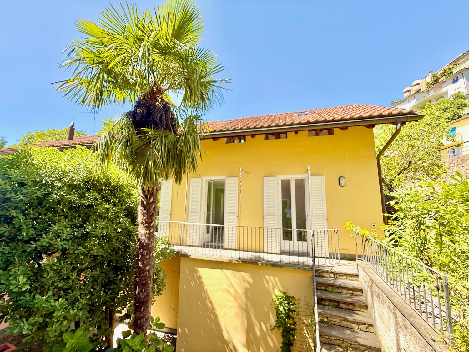 Two-story house, yellow exterior, red tile roof, balcony on the first floor, windows with white shutters, steps leading up to the entrance, front yard with palm tree and greenery.