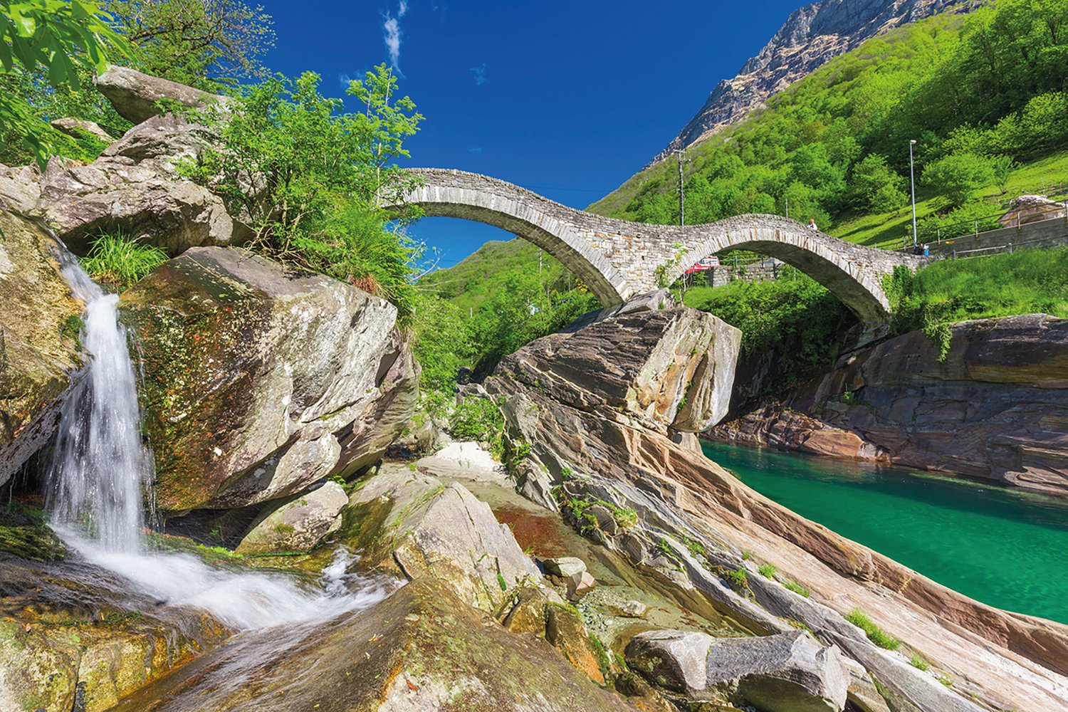 stream flowing through rocky landscape, bridge across the water, waterfall, lush greenery, mountain in the background