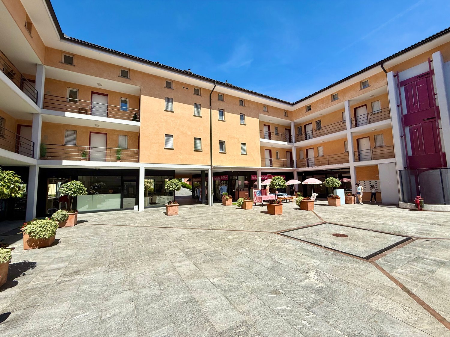 Modern multi-story building with a paved courtyard, balconies, multiple entrances, and trees.