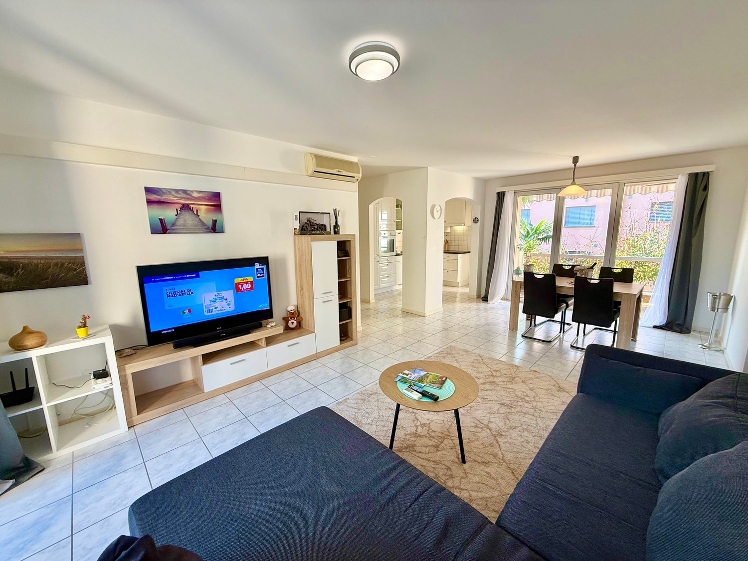 Spacious room with a flat-screen TV, a large blue sofa, a wooden table, a dining table, white tiled floors, and glass doors leading to an outdoor balcony