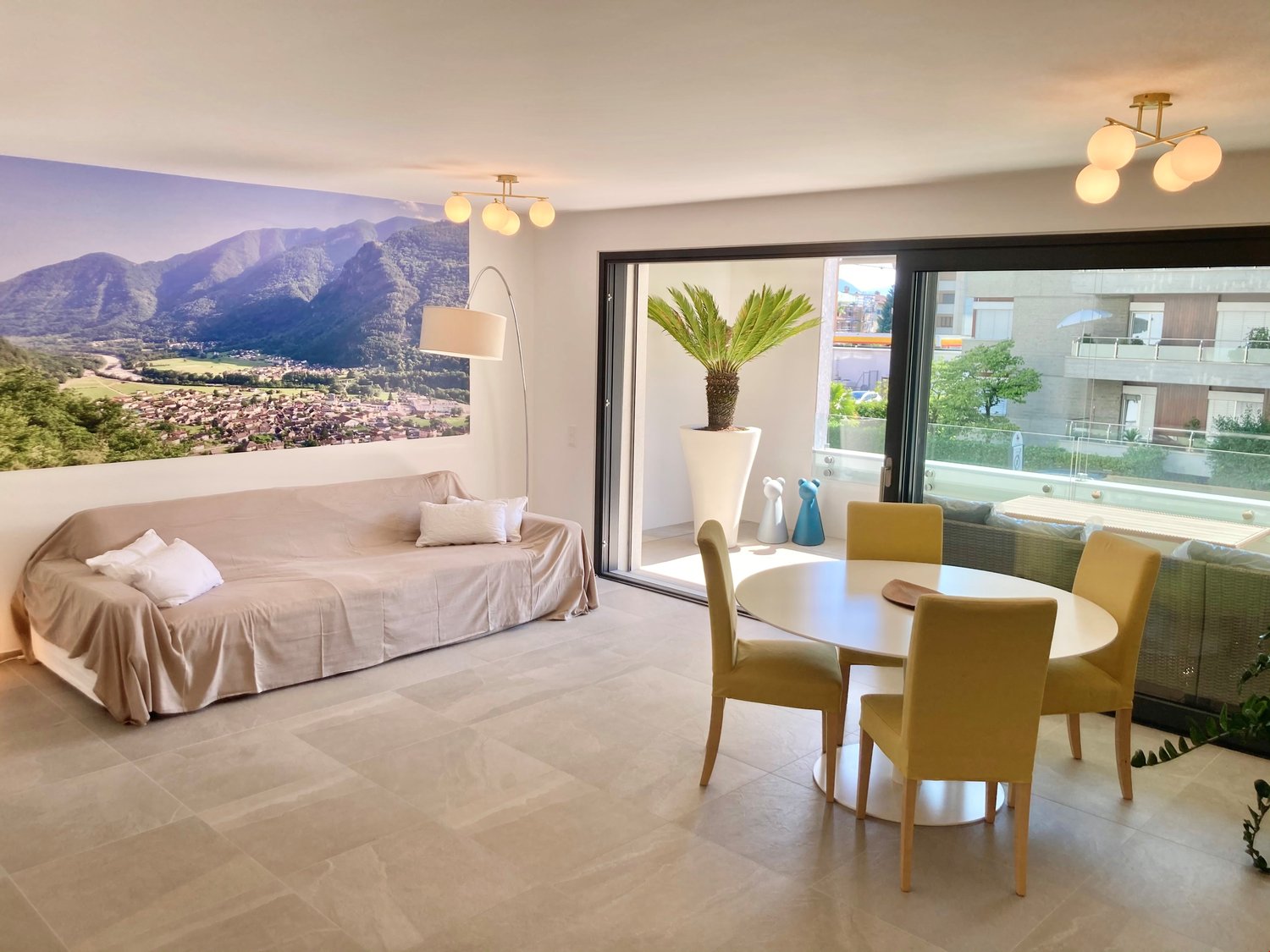 Living room with beige sofa, lamp, dining table with chairs, sliding glass door to balcony, mountain view wallpaper.