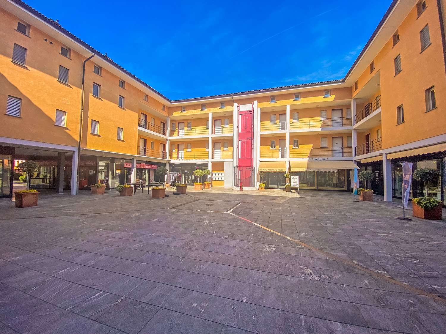 yellow apartment building, glass storefronts, multiple balconies, central courtyard with stone pavement and potted plants