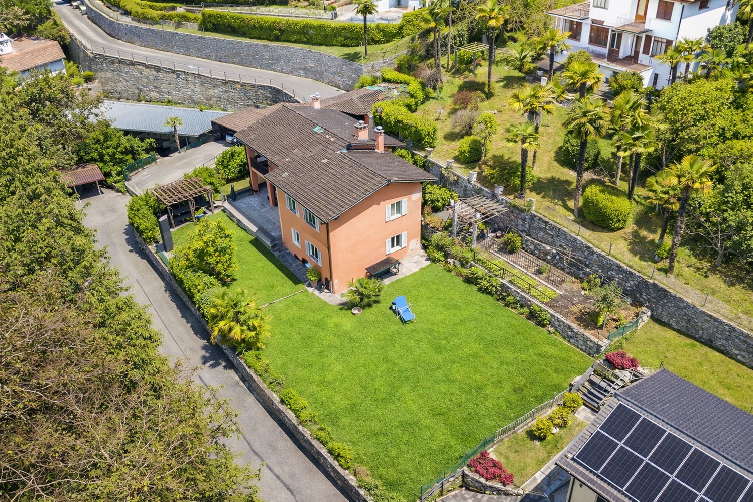 Large 2-story house, brown roof, garden, palm trees, large grass, lounge chair, solar panels on roof