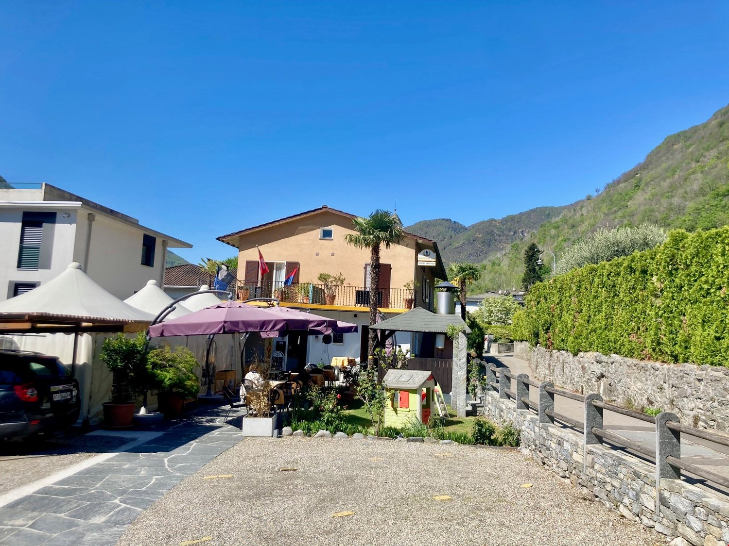 A building with two floors, beige exterior, outdoor dining tables, plants, flags, a small house, mountains, and a clear sky.