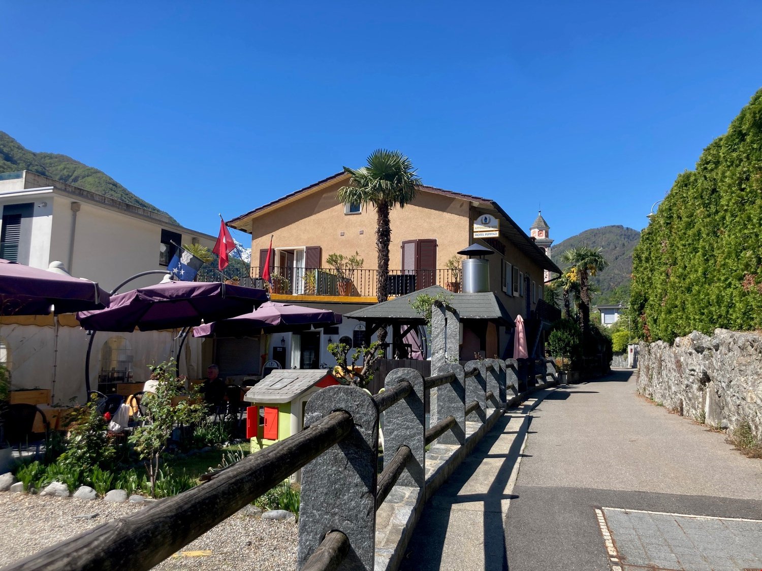 2 story hotel with outdoor dining area, flags, red awnings, balcony with plants, palm trees, stone fence, concrete walkway, mountain in background
