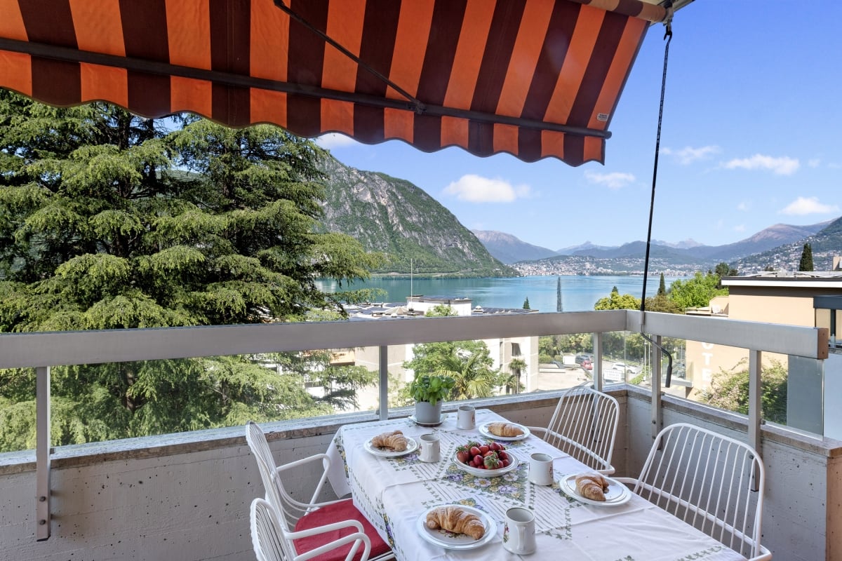 Balcony with a view of a lake surrounded by mountains, with a table set for breakfast or brunch with croissants, fruit, and other items.