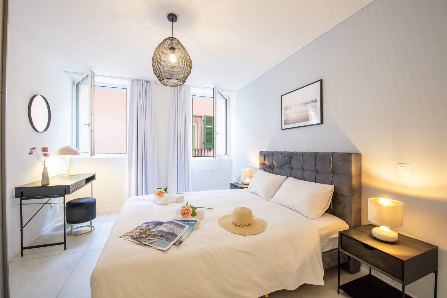 King size bed, white walls, two nightstands with lamps, desk with round mirror, round hanging lamp, window with balcony, picture frame