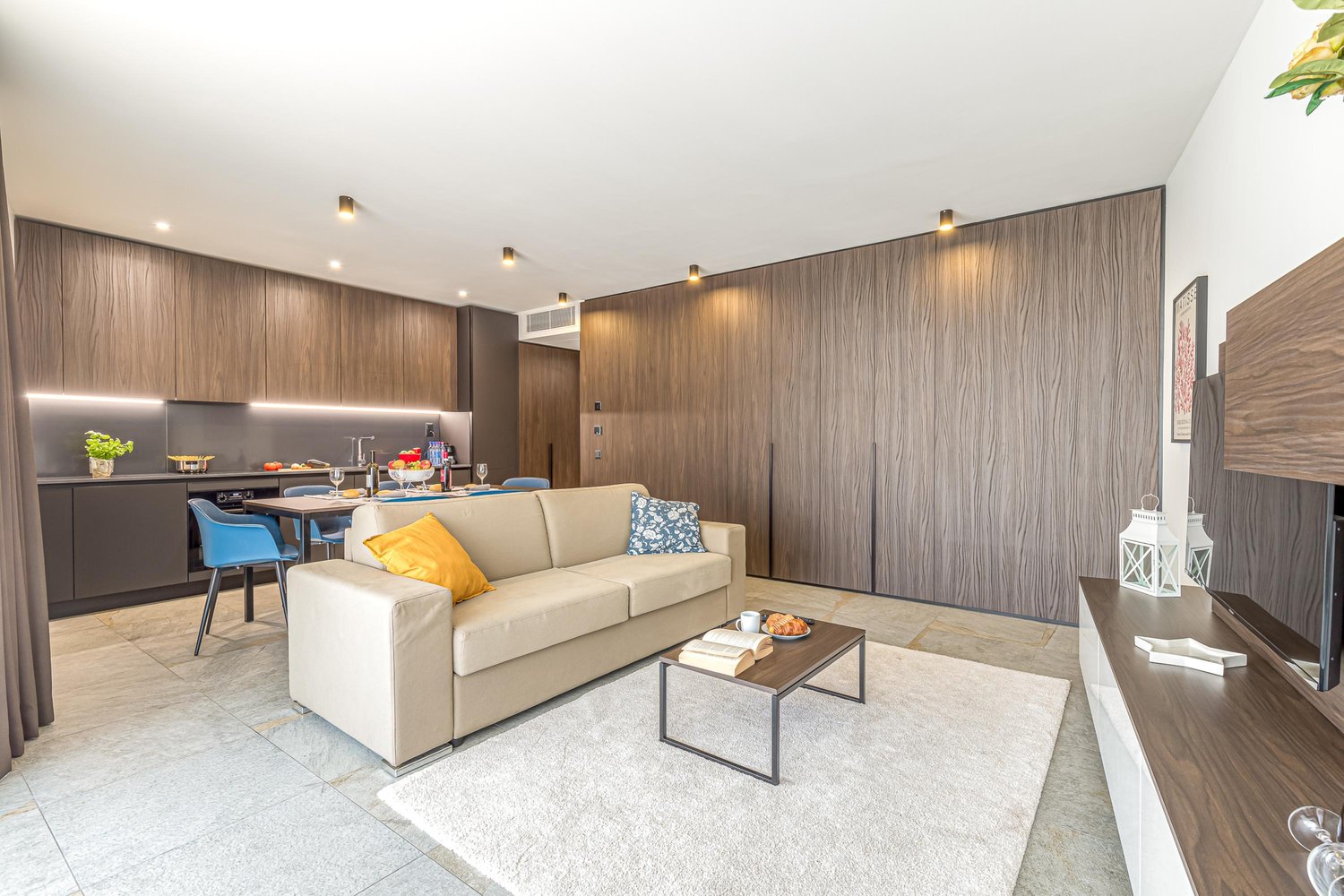 Modern living area with beige couch, white rug, coffee table, TV unit, dining table with chairs