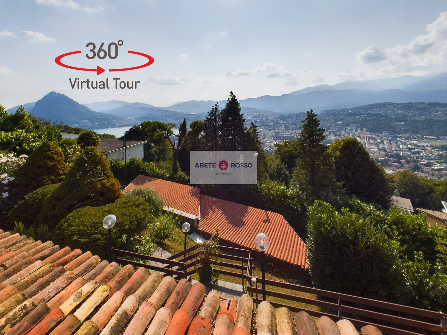 The image shows a panoramic view of a mountainous landscape with a city in the distance. The foreground features a building with a red tile roof and the Abete Rosso real estate logo. The surrounding area is lush with trees and greenery, providing a scenic