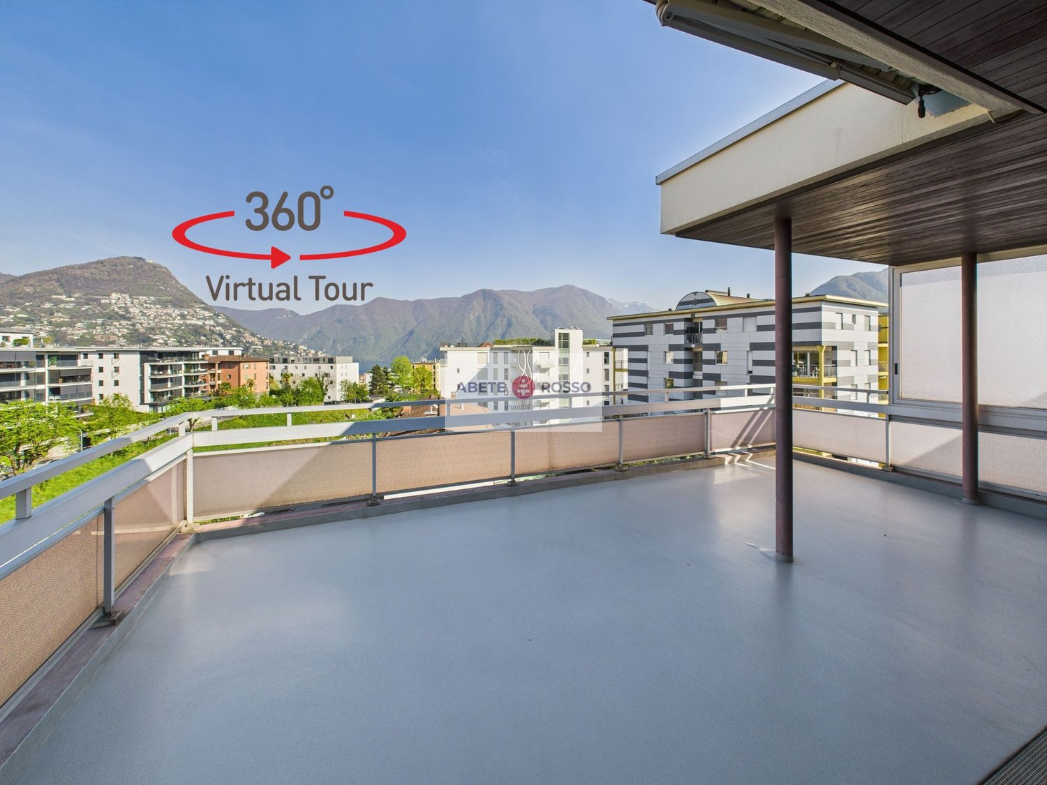 The image shows a 360-degree virtual tour of a balcony or terrace overlooking a city. The balcony has a concrete floor and a wooden roof structure. The view includes several multi-story buildings in the distance, with mountains visible in the background.