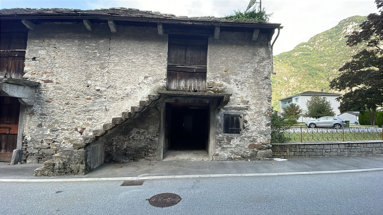 Two-story stone house, wooden staircase, dark entrance, stone walls, two cars parked on the road