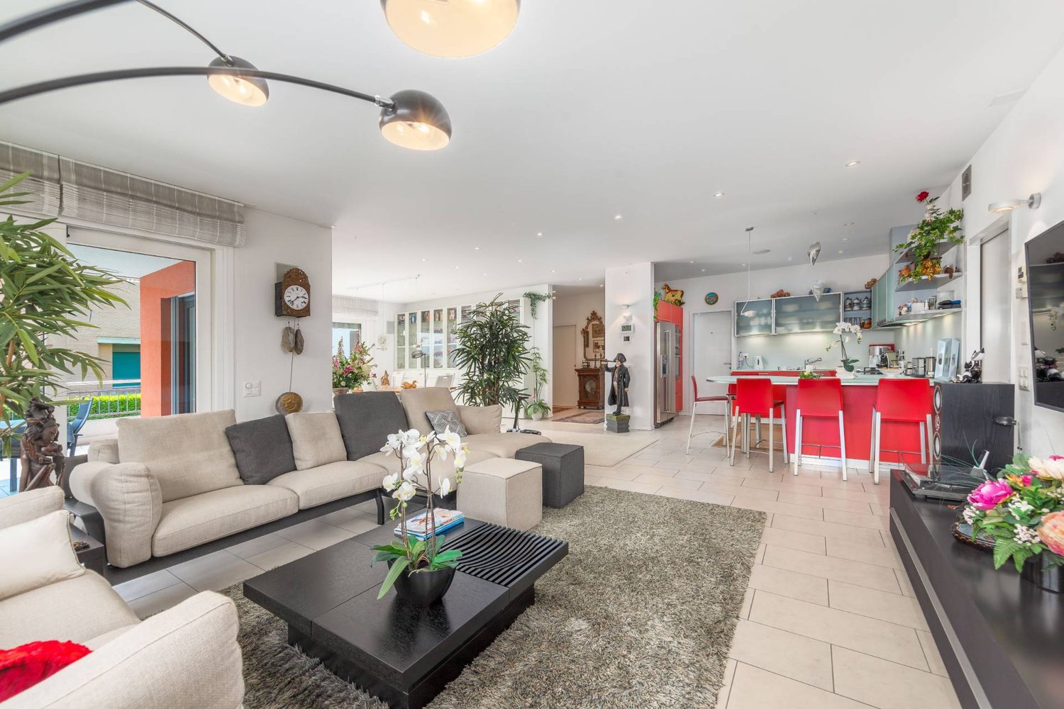 Open-concept living room and kitchen, with a large sofa, coffee table, and dining area. The room has large windows, track lighting, and decorative plants. The kitchen features red chairs and modern appliances.