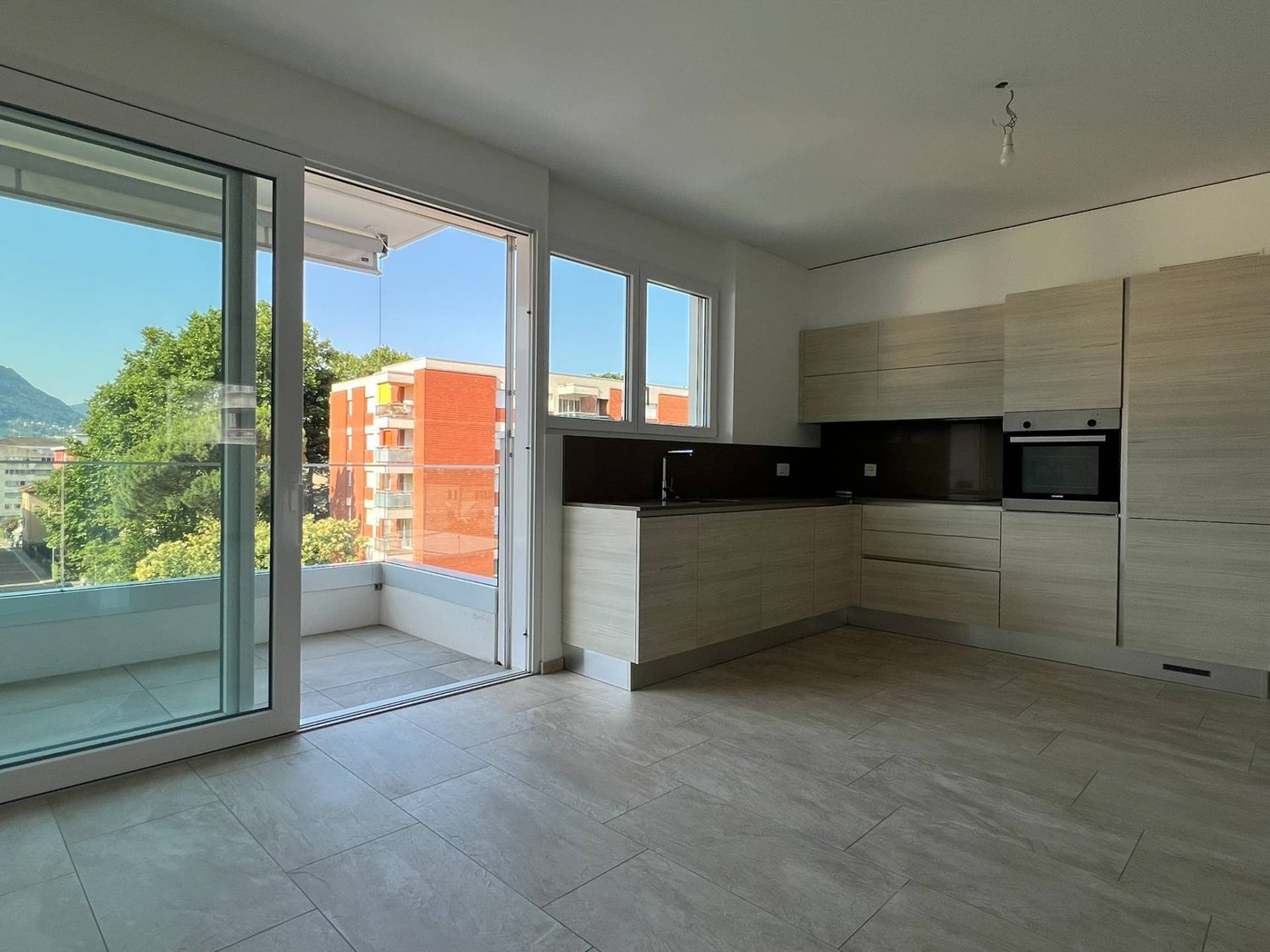 Spacious open-plan living area with large windows and sliding glass doors leading to a balcony. The kitchen features modern wood-toned cabinets and appliances. The flooring is a neutral-colored tile throughout the space.