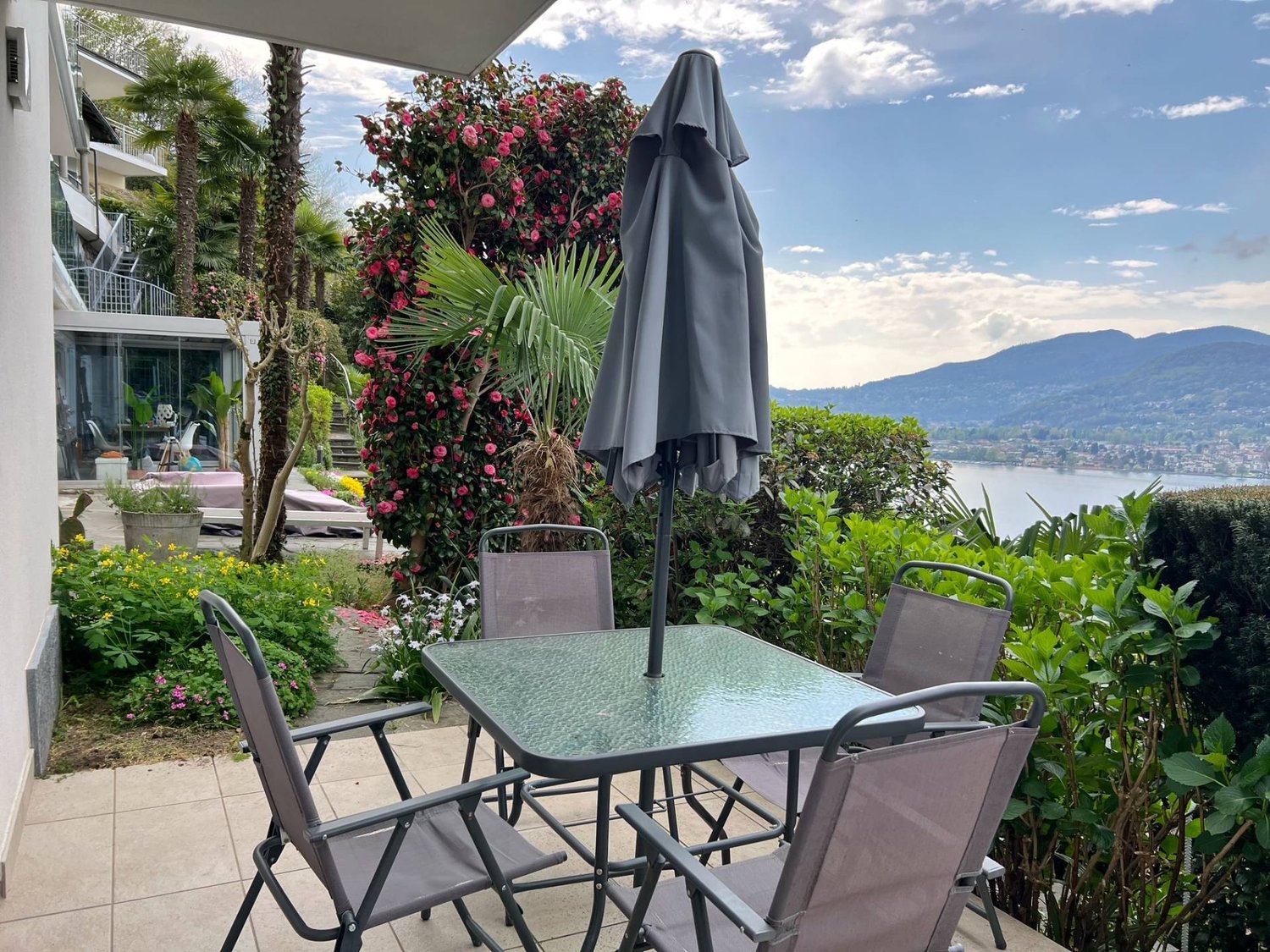 Outdoor patio with a glass-topped table and chairs, surrounded by lush tropical plants and flowers. There is a closed umbrella and a view of the mountains and lake in the background.
