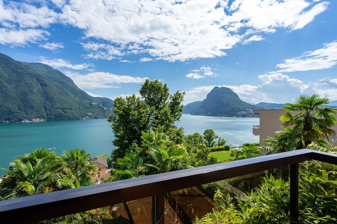 The image shows a scenic view of a lake surrounded by mountains and lush greenery. There is a balcony or terrace in the foreground, with palm trees and other tropical vegetation visible. The sky is blue with fluffy white clouds, creating a picturesque and