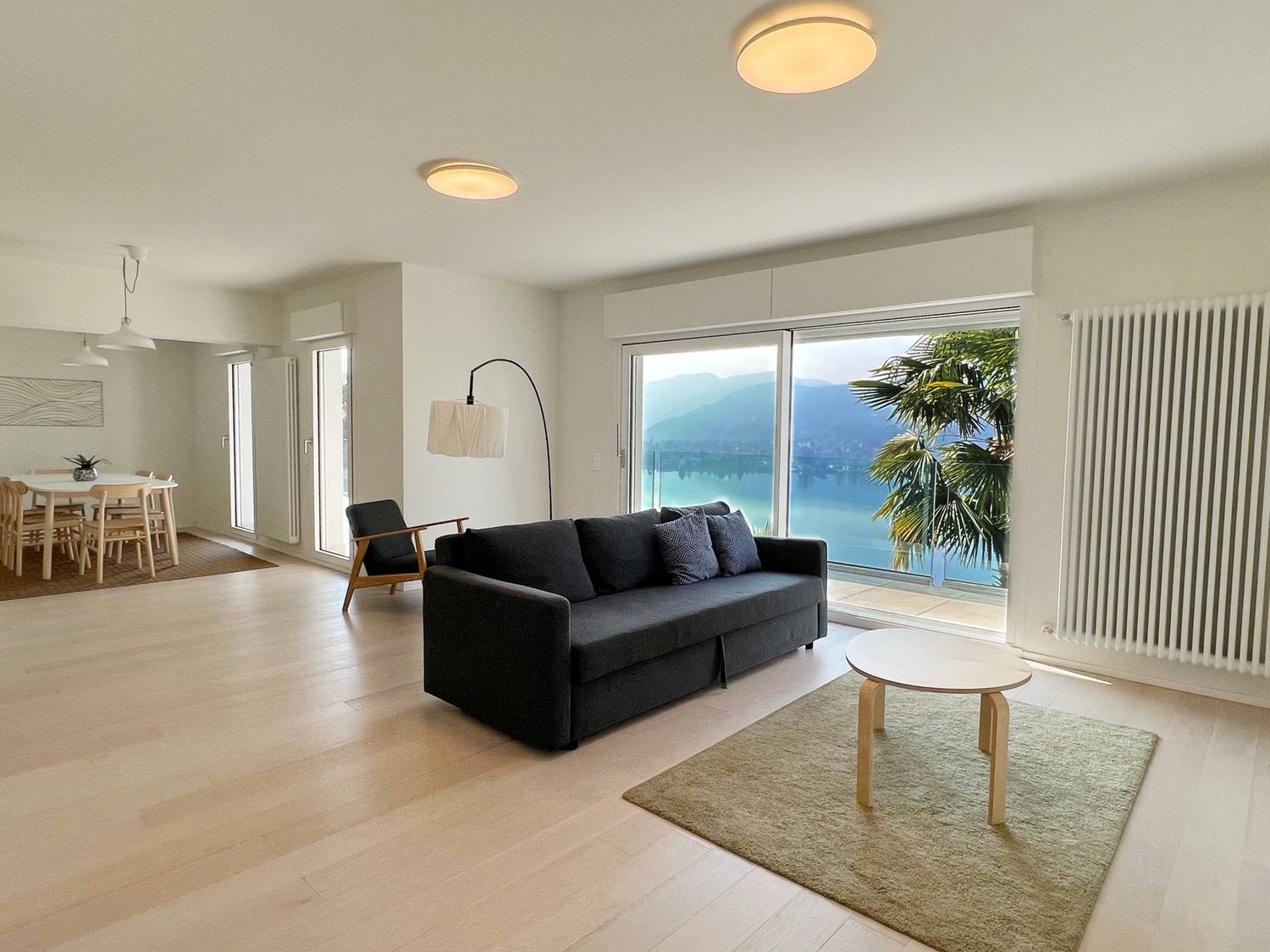 Spacious living room with large windows offering a scenic view of the lake and mountains, hardwood floors, black sofa, wooden coffee table, and a potted plant adding a touch of greenery to the space.