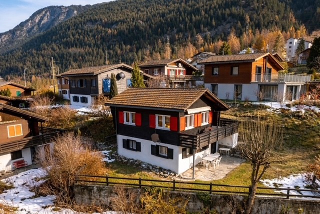 Mountain chalet, brown roof, red shutters, balcony, surrounded by other chalets, snowy mountain in the background