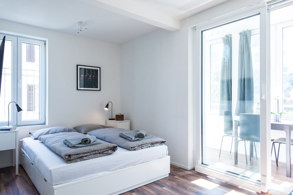 Spacious bedroom with large windows, white walls, hardwood floor, bed with gray bedding, nightstand, and artwork on the wall.