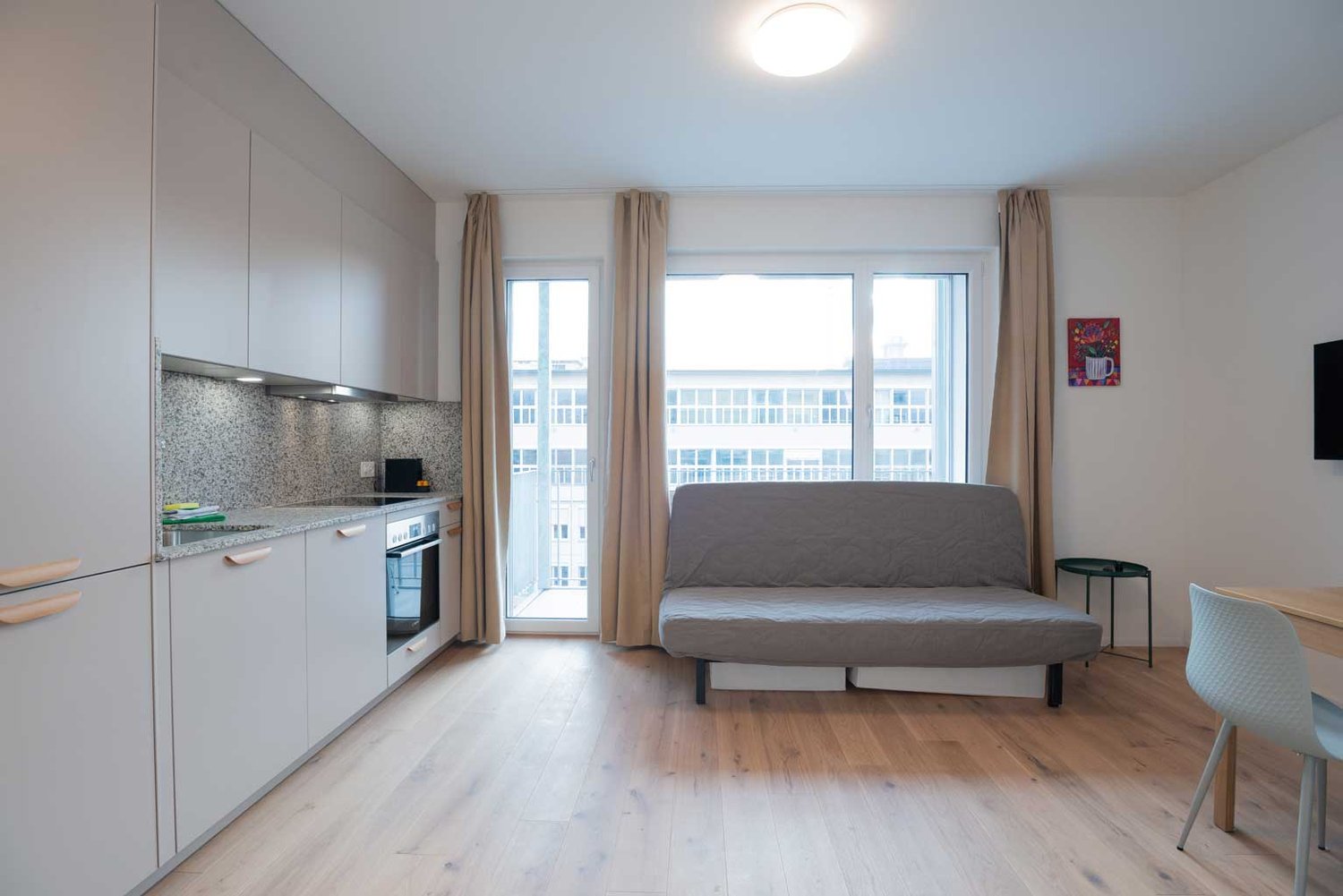 Open-plan studio apartment with kitchen, living area, and balcony. The apartment features hardwood floors, large windows, white walls, and modern kitchen cabinets. The living area has a gray sofa bed and a small side table. The balcony is accessible throu