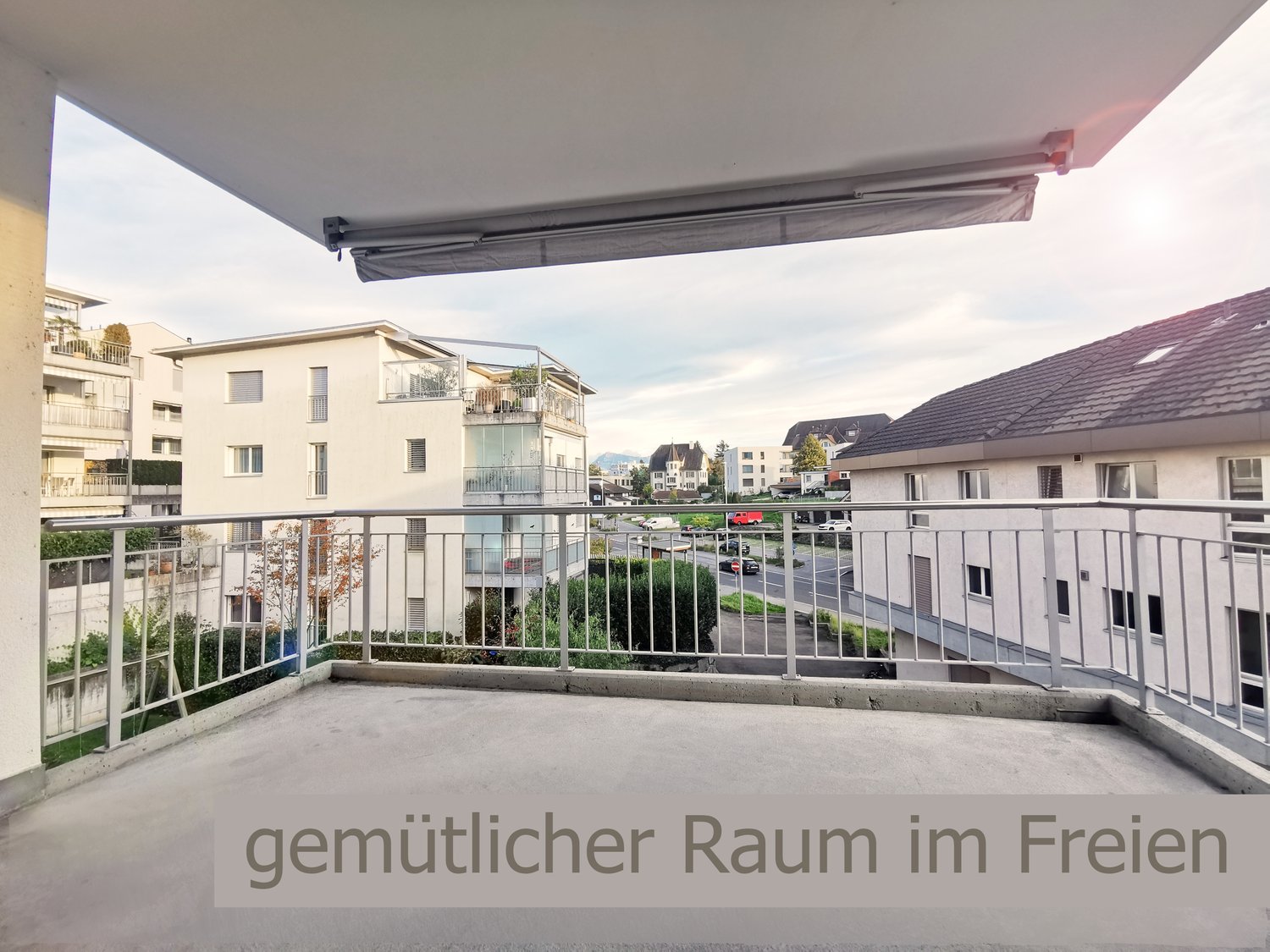 The image shows a balcony or terrace with a retractable awning. The balcony overlooks a residential area with several multi-story apartment buildings. The text at the bottom of the image reads 'gemutlicher Raum im Freien', which translates to 'cozy space