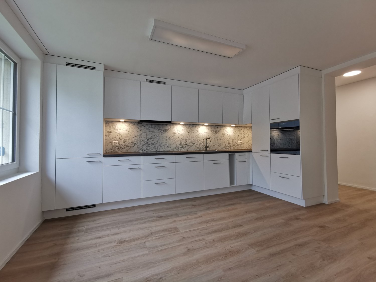 White wooden cabinets, light wooden floor, ceiling with recessed lighting, window to the left