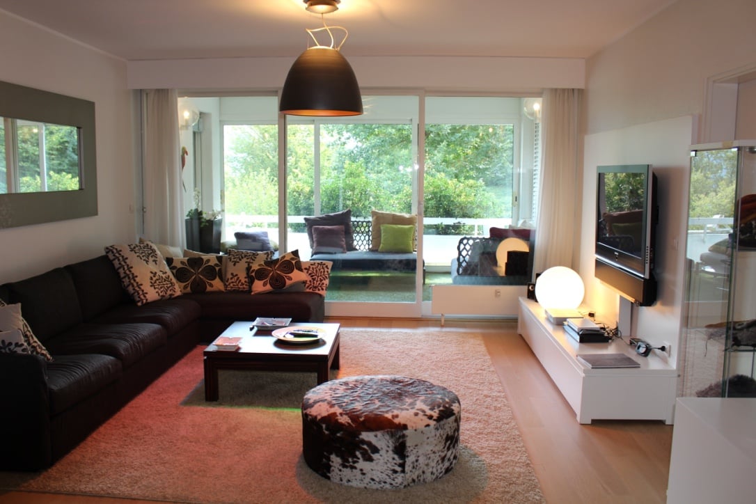 Spacious living room with large windows overlooking a balcony or terrace, dark leather sofa, patterned throw pillows, coffee table, and entertainment unit with a TV.