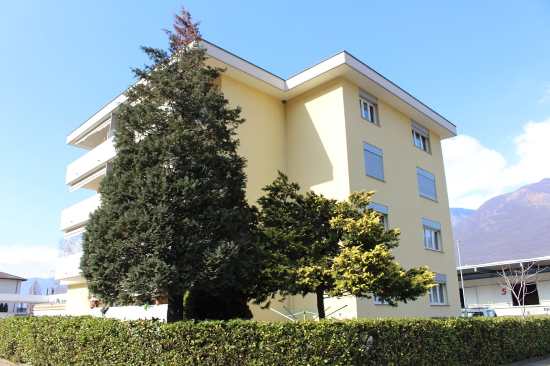 3-story apartment building with yellow exterior, large windows, balconies, and surrounded by trees and shrubs
