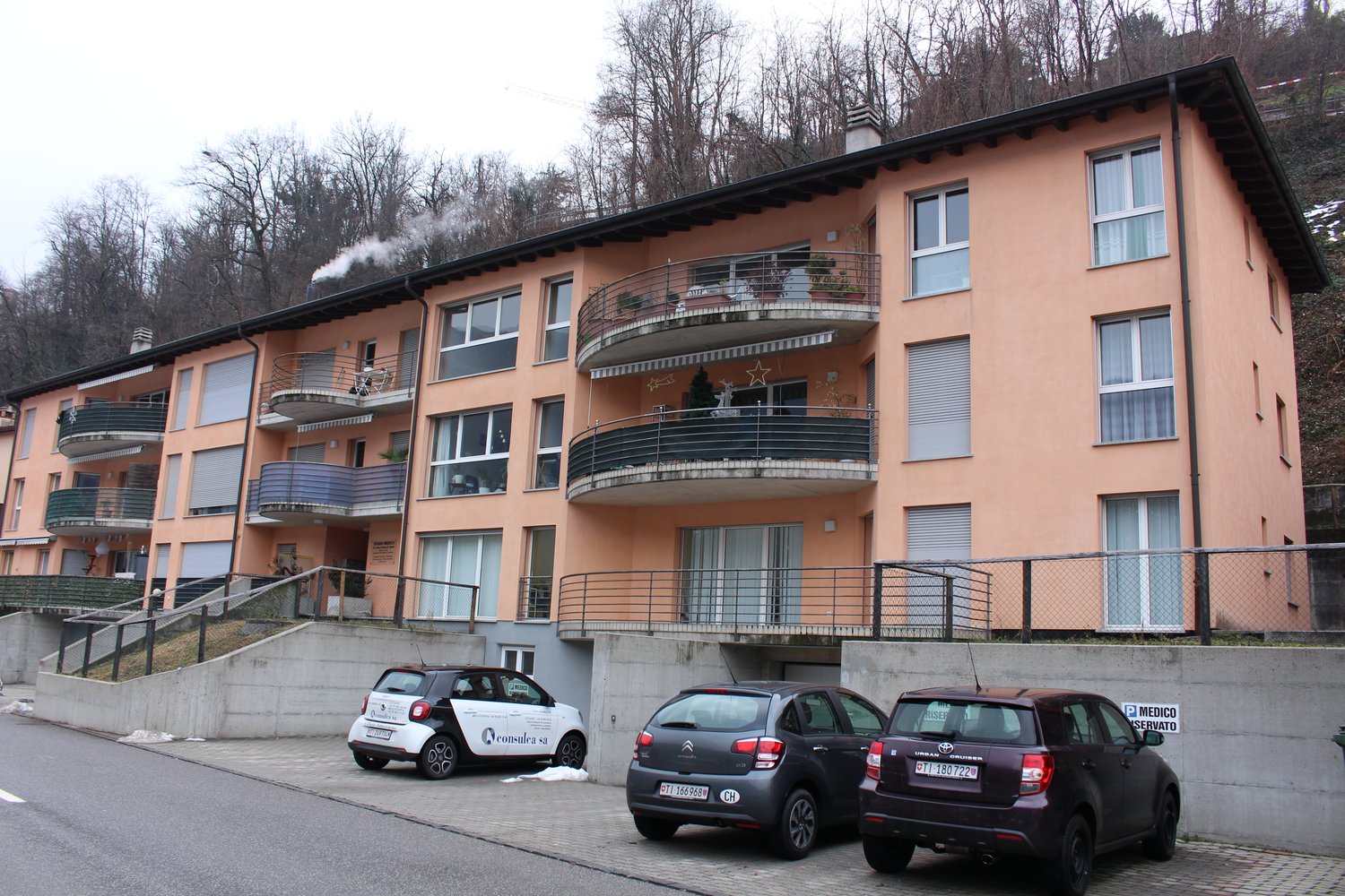 Multi-story building with balconies, multiple cars parked in front, beige exterior, large windows, staircase leading to entrance, fenced-in area