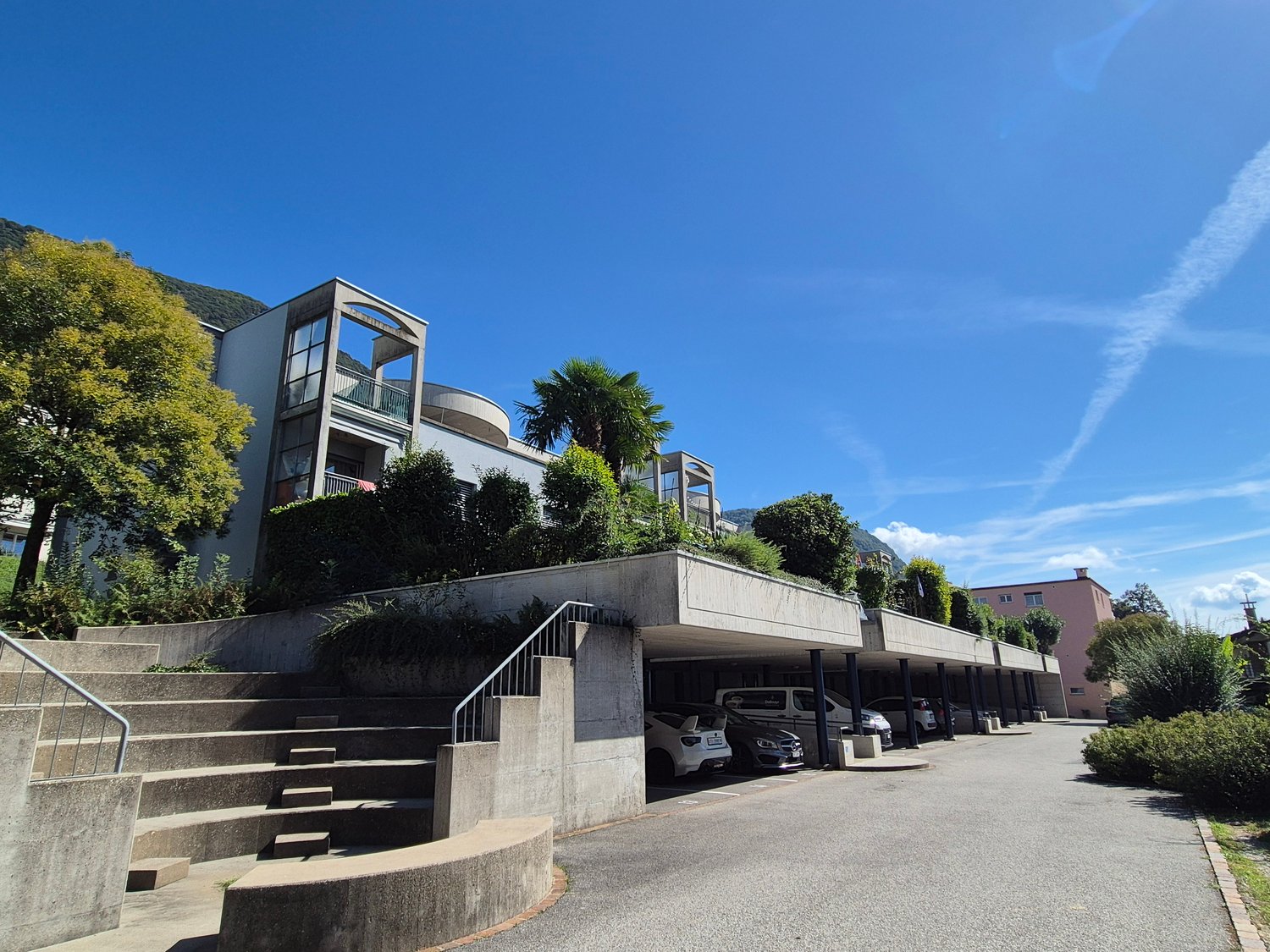 Modern house, large balcony on the top floor, multiple cars parked in underground parking, stairs leading to parking