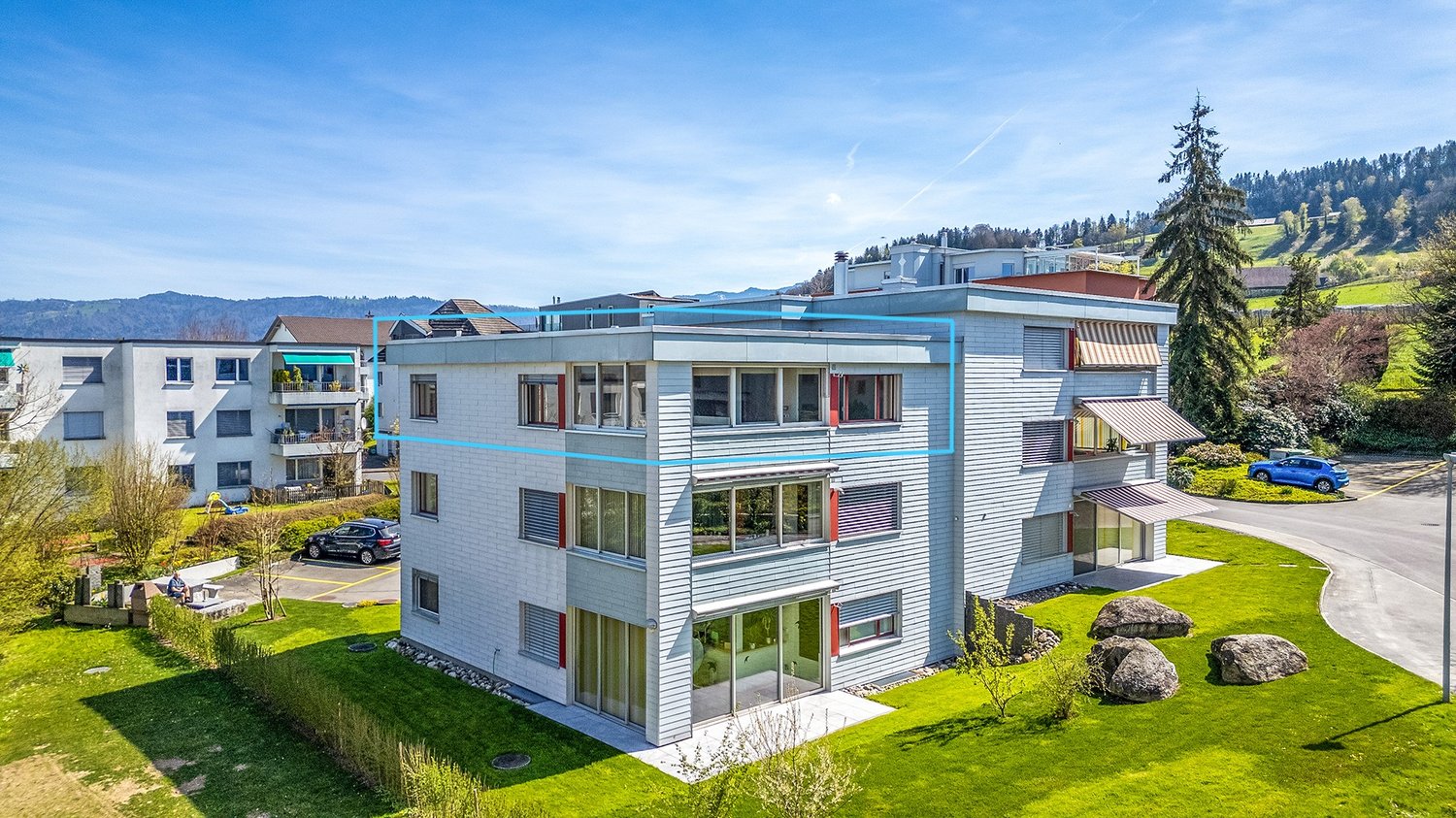 3-story modern apartment building with white and blue exterior, large windows, balconies, and a parking area in the foreground. The building is surrounded by lush greenery and trees, with mountains visible in the background.