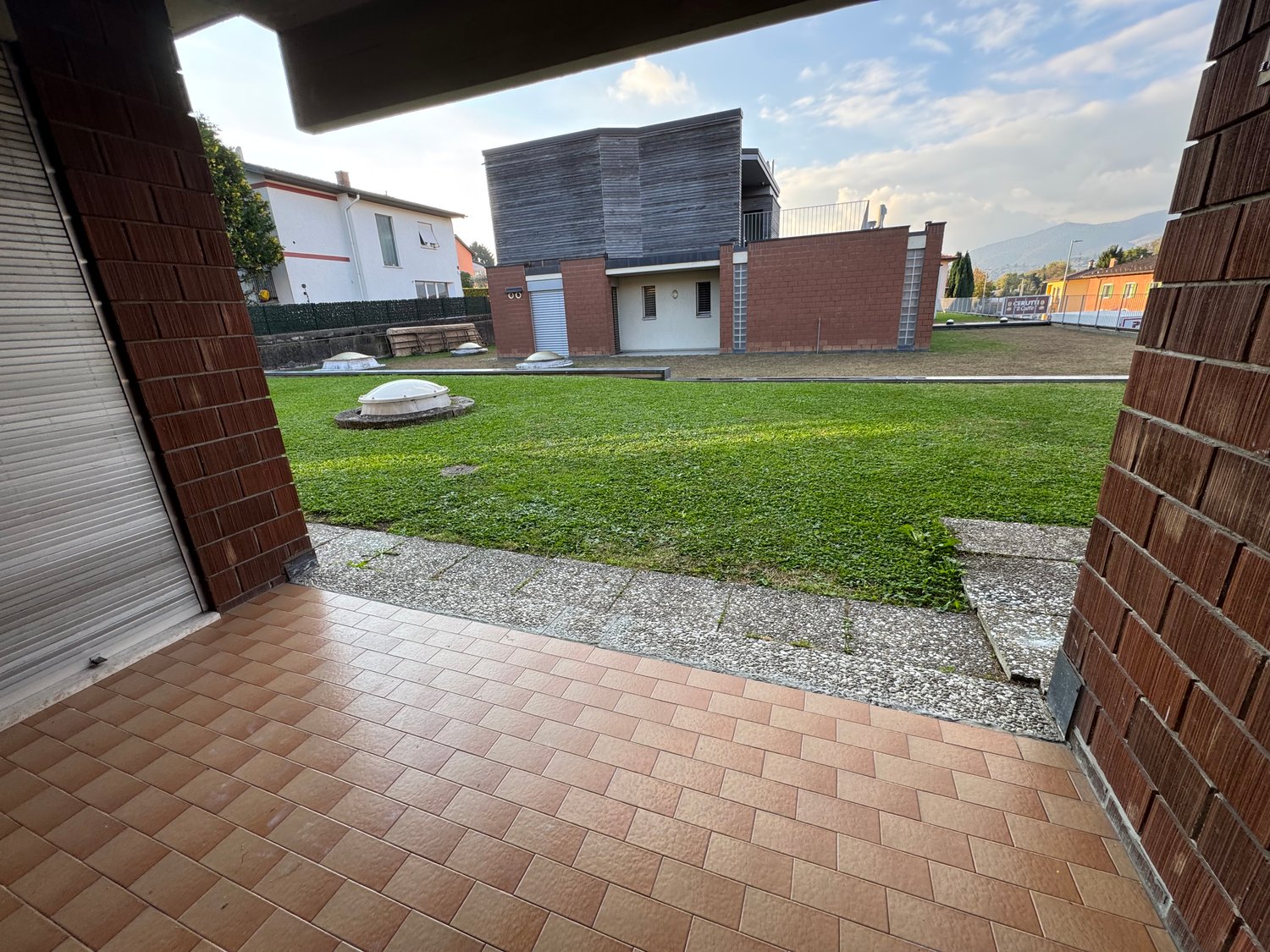 A backyard area with a brick floor and a spacious green lawn, surrounded by buildings and trees. The area has a clear sky with some clouds.