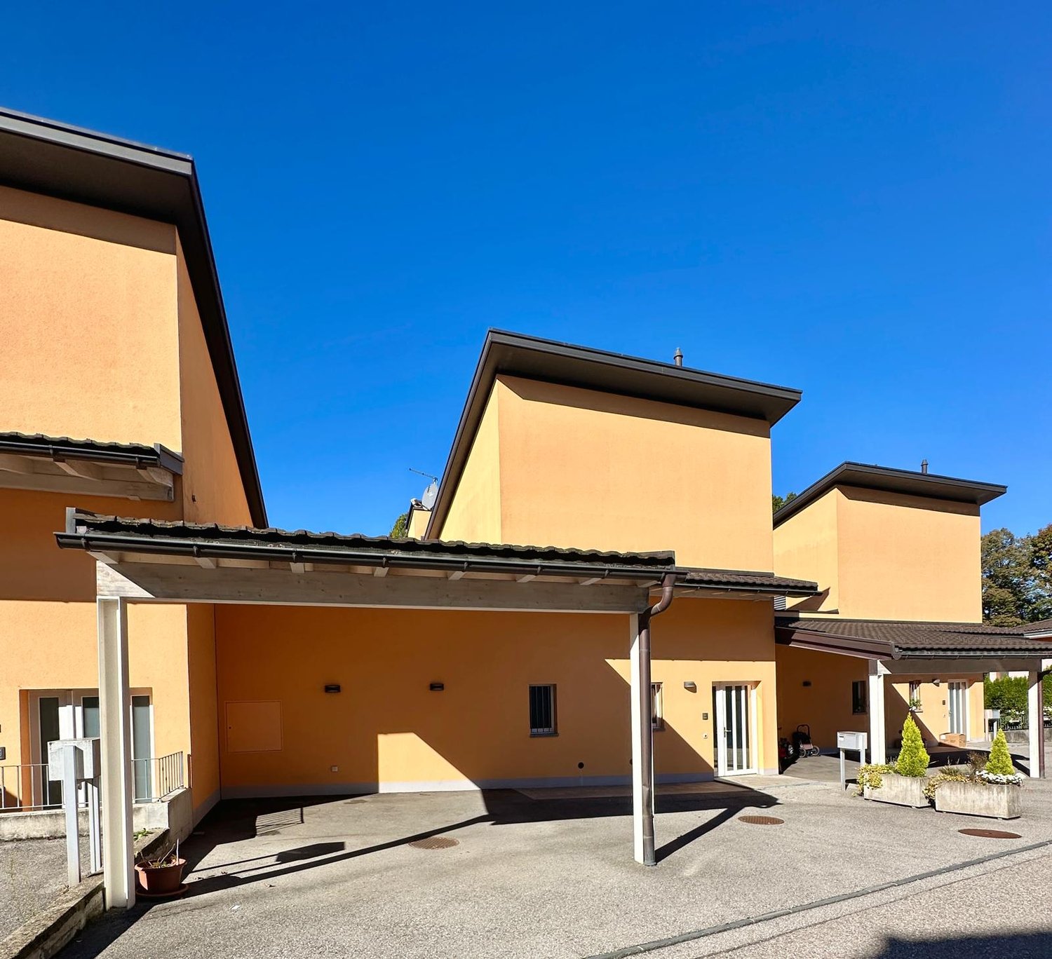 Three-story building, cream colored walls, flat roof, central paved courtyard, large windows, small balconies on second floor, several large potted plants.