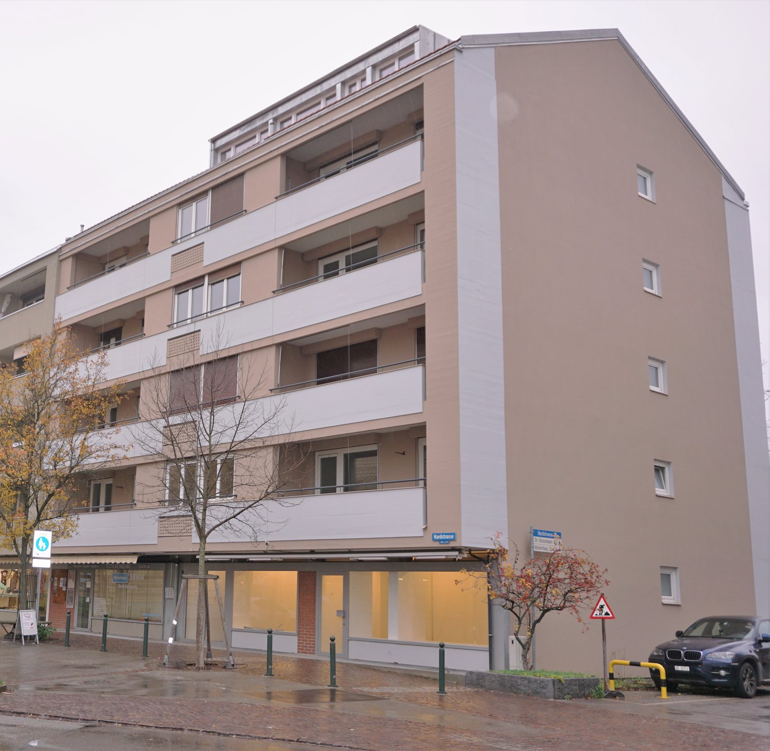 An apartment building with multiple balconies, beige exterior, glass doors and windows, and several trees in front.