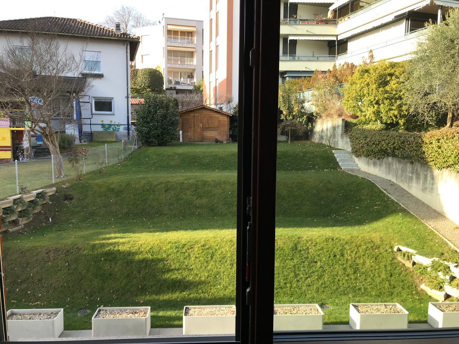 The image shows a view from an apartment building, with a grassy yard and garden area in the foreground. There are several apartment buildings visible in the background, and a small wooden shed can be seen in the yard. The apartment building has balconies