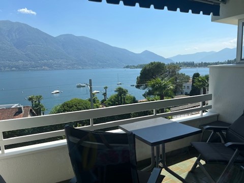 Balcony overlooking a lake surrounded by mountains, with a table and chairs set up for outdoor seating.