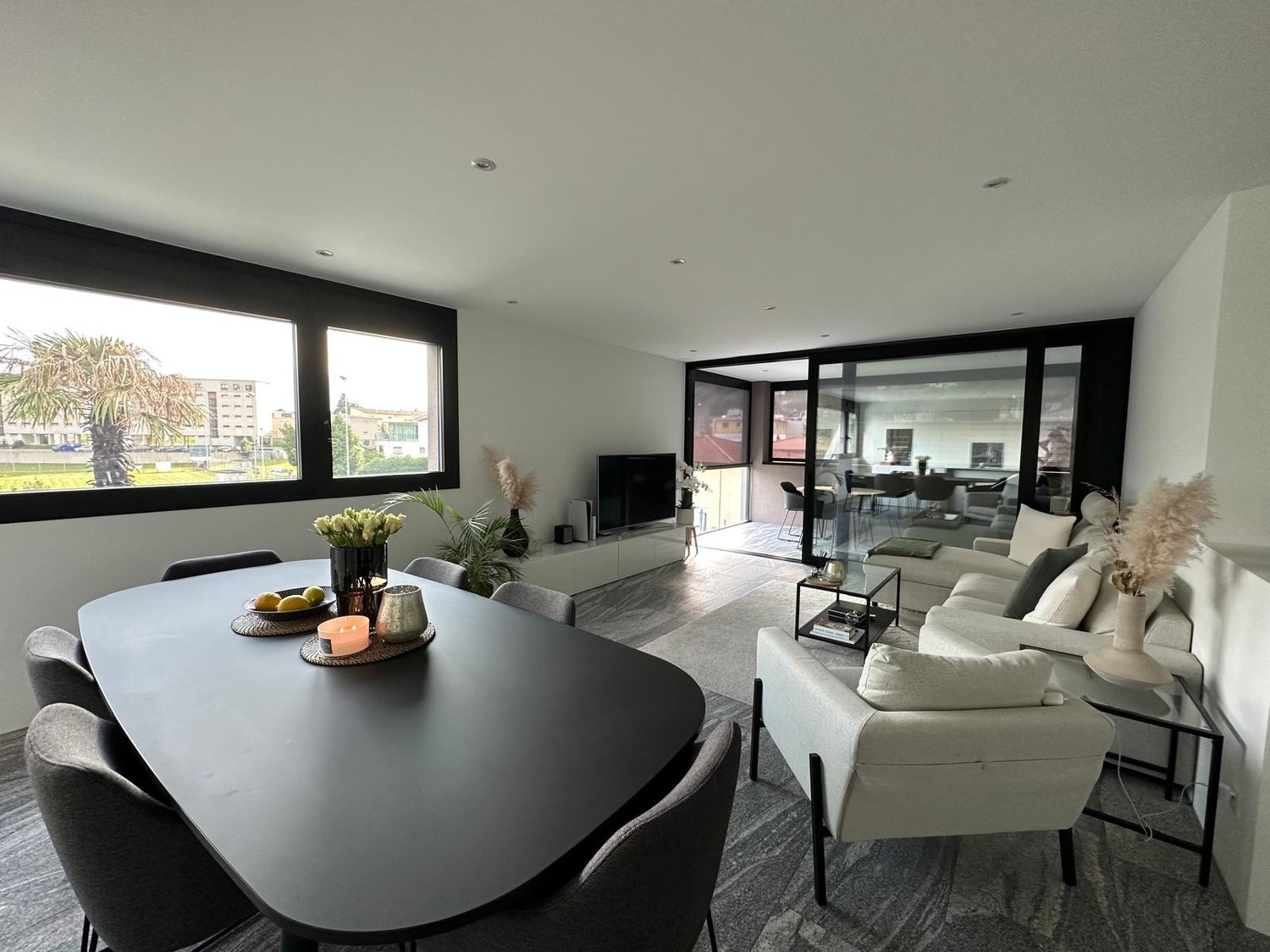 A modern, minimalist living room with a dining table and chairs, a sofa, a TV, and glass doors leading to a balcony.