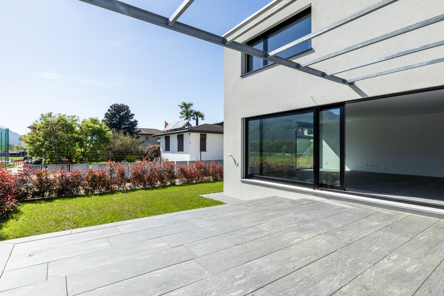 modern house with gray tiled terrace, large glass doors and windows, greenery in the background